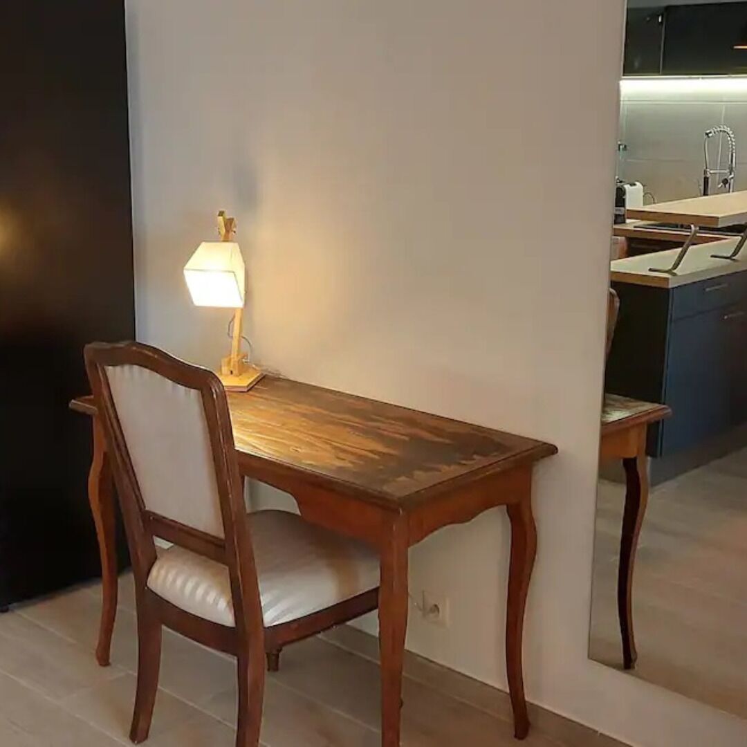 A dining area with a wooden table and chair, a table lamp, and a kitchen in the background.