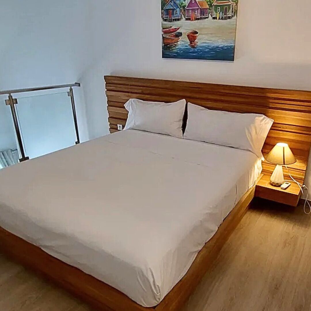 Un lit bien fait, avec une tête de lit en bois, une table de chevet avec une lampe et un tableau représentant des maisons accroché au mur.