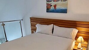 Een net opgemaakt bed met een houten hoofdbord, een nachtkastje met een lamp en een schilderij van huizen aan de muur.