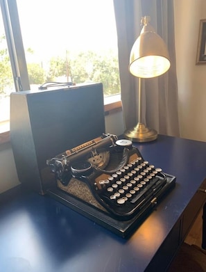 Typewriter sits atop a desk in front of a window, accompanied by a desk lamp.