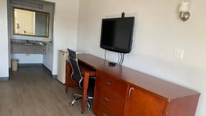 A compact room with a wooden desk, a black monitor, and a small kitchenette in the background. - American Inn & Suites (Forest Park)