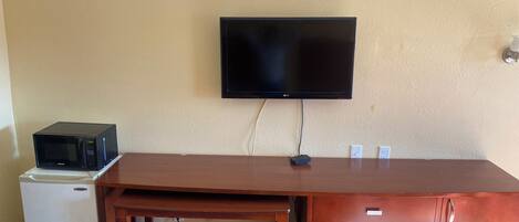 Una habitación con un escritorio de madera, un televisor montado en la pared, un microondas y un refrigerador.