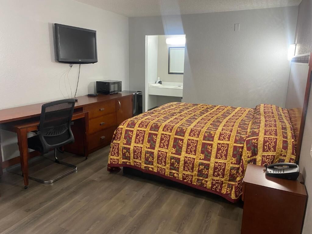 Habitación de hotel con cama, escritorio con silla, televisor, microondas y una pequeña mesa con control remoto.