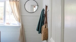 A room with a window, white wooden walls, a round mirror, and a coat rack with a green jacket and a woven bag.