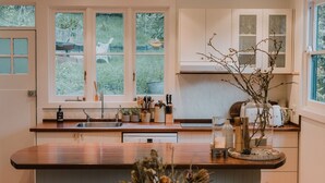 A modern kitchen with a wooden dining table, a hanging light fixture, and a view of the garden through the window.