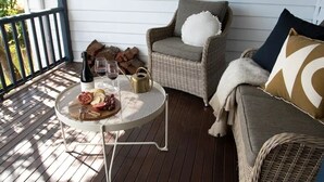 A wicker chair with a white cushion and a throw blanket, a small round table with a bottle, glasses, and food, and a wooden deck with a railing.