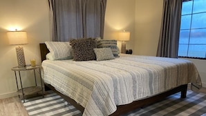A neatly made bed with a striped bedspread, two bedside tables with lamps, a checkered rug, and a window with curtains.
