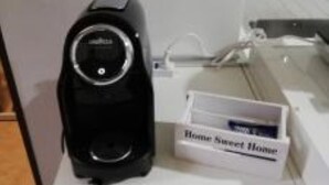 Une machine à café noire est posée sur une table blanche, à côté d’une boîte sur laquelle est écrit « Chez soi, quel bonheur ! ».