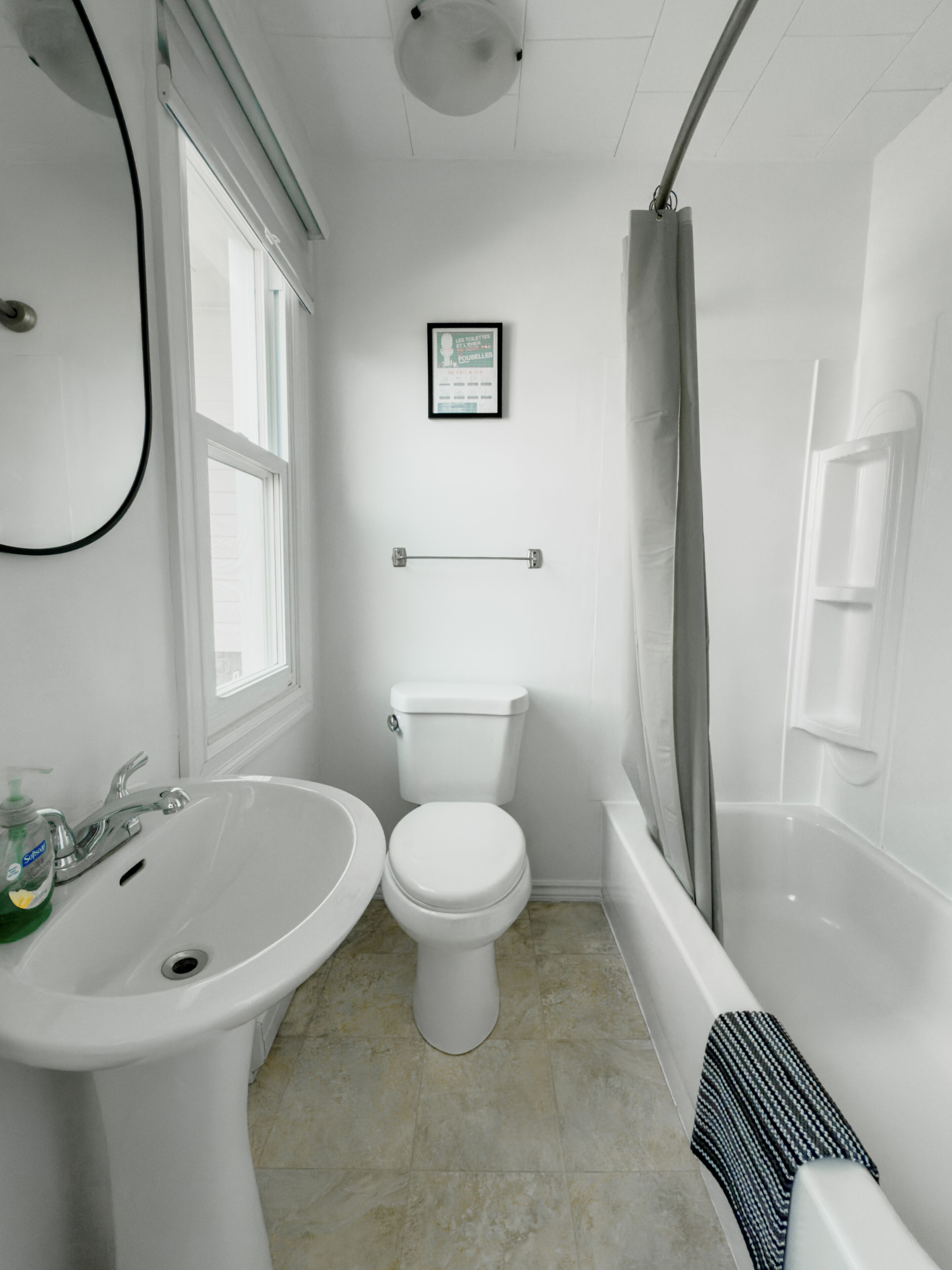 Une salle de bain avec un lavabo, des toilettes et une baignoire, tous de couleur blanche. Il y a une fenêtre sur la gauche et un porte-serviettes au-dessus des toilettes.