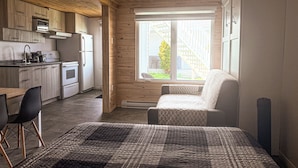 Habitación compacta con techo de madera, kitchenette, un ventanal grande y cama con manta estampada.