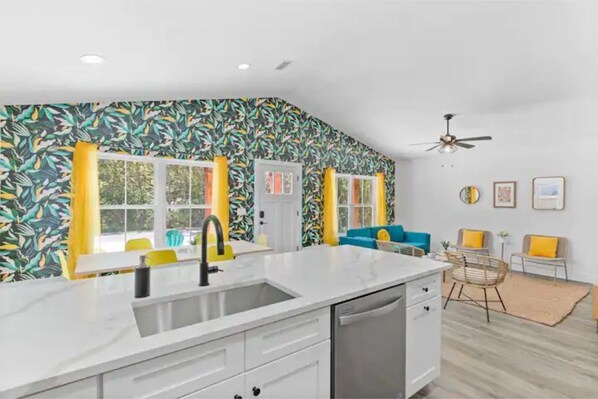 A modern kitchen with a white countertop, stainless steel sink, and a built-in dishwasher. There are yellow curtains on the windows, and a ceiling fan is visible.
