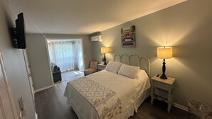 A bedroom with a bed, bedside lamps, a chair, a window with curtains, and a wall-mounted air conditioner. - Room 2 - The Inn at Dirickson Creek (Frankford)