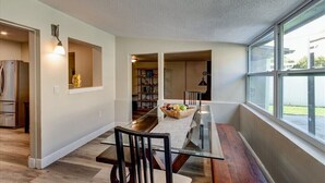 A modern kitchen with a dining area, featuring a glass table, wooden chairs, and a large window providing natural light.