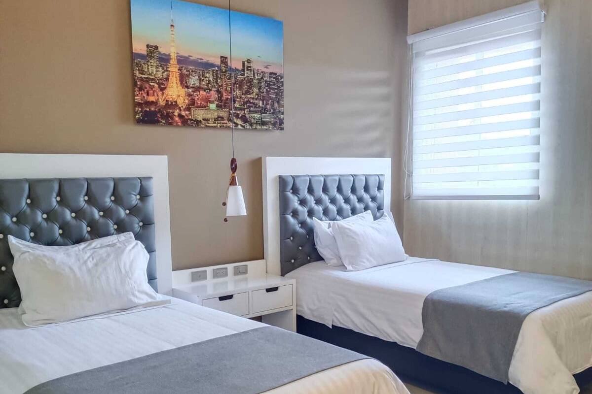A hotel room with two beds, a cityscape painting, a window with blinds, and a nightstand.