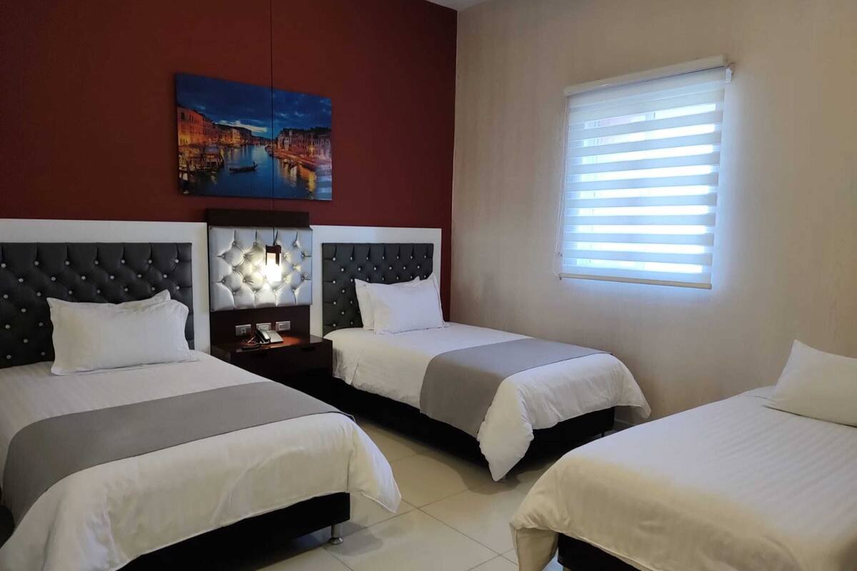 A hotel room with three beds, a painting on the wall, a nightstand with a lamp, and a window with blinds.