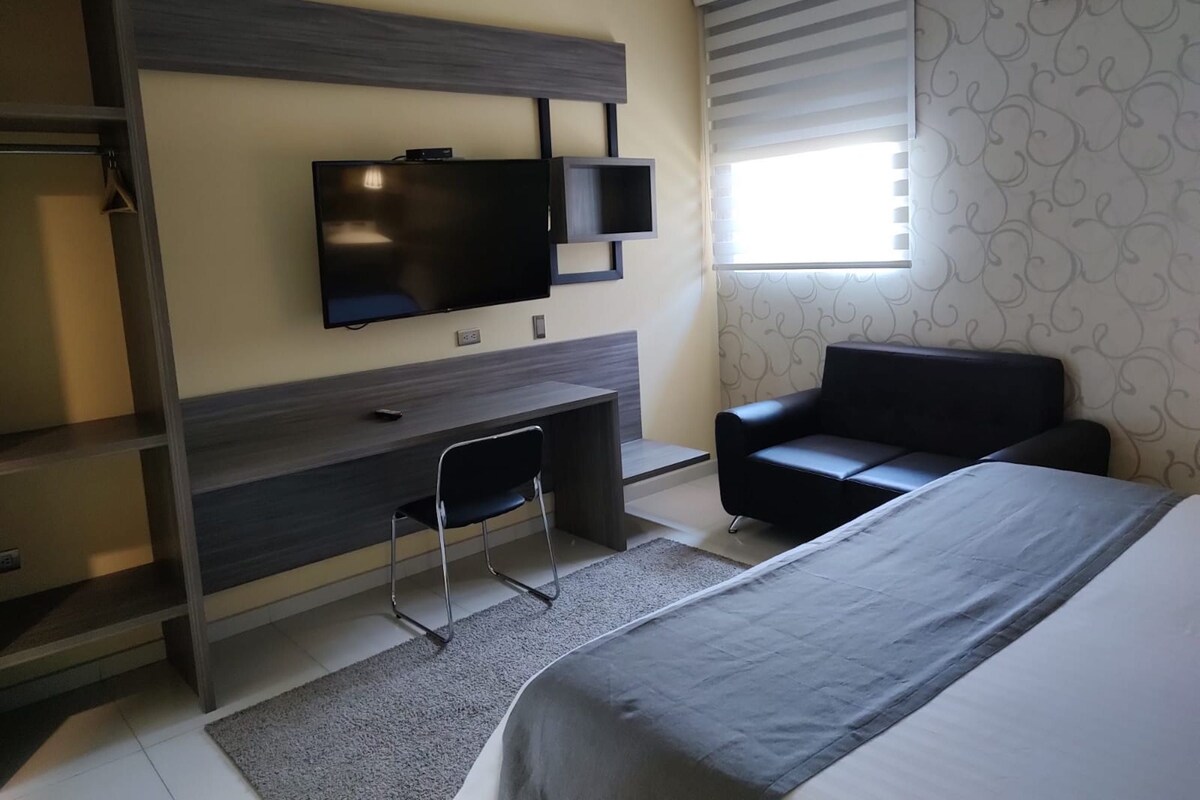 A hotel room with a bed, a desk, a chair, a TV, and a window with blinds.