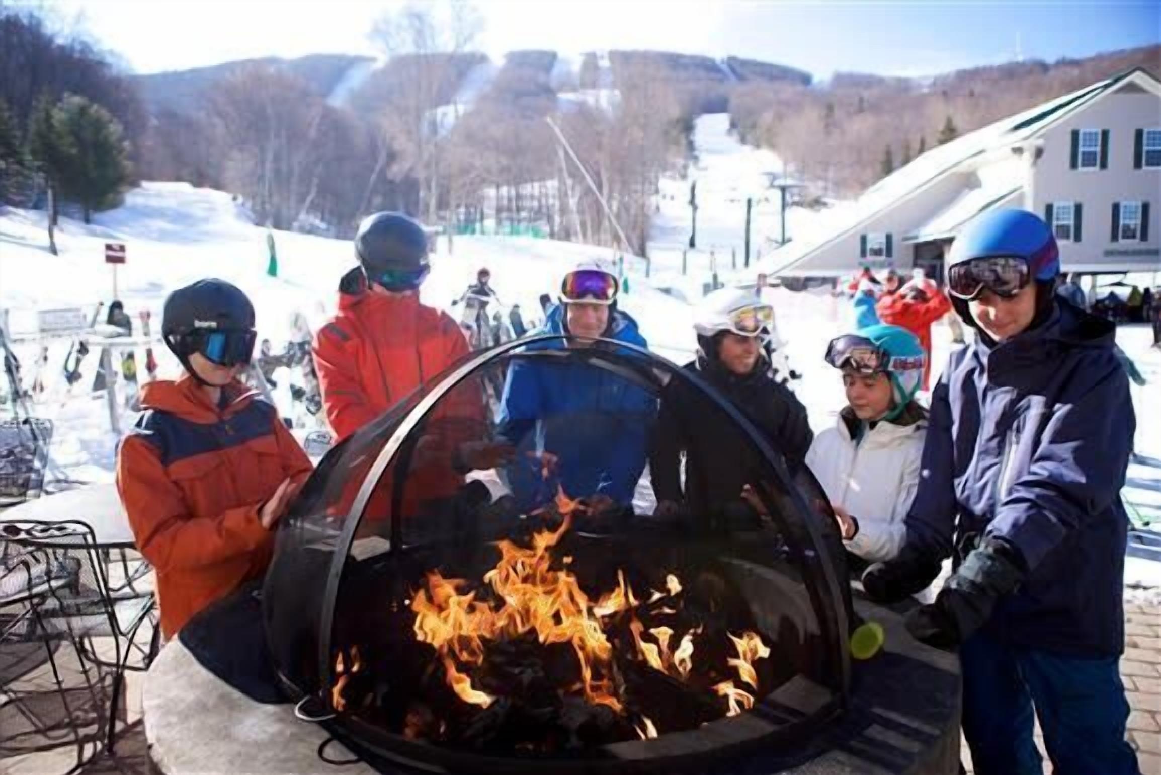 A group of people around a glass enclosure with a fire inside, outdoors in a snowy setting.