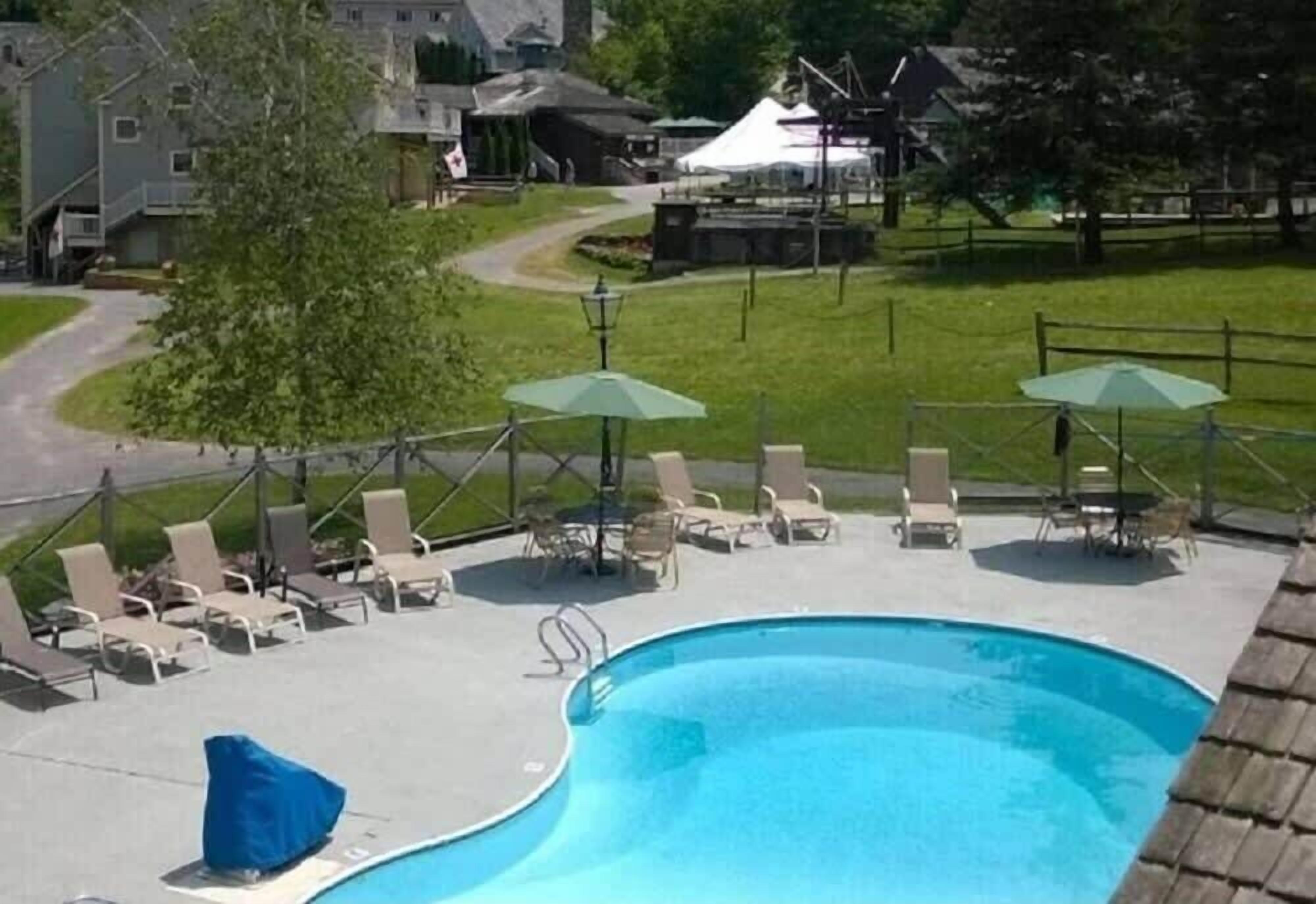 En swimmingpool med liggestole og parasoller, omgivet af et hegn og boligbygninger.