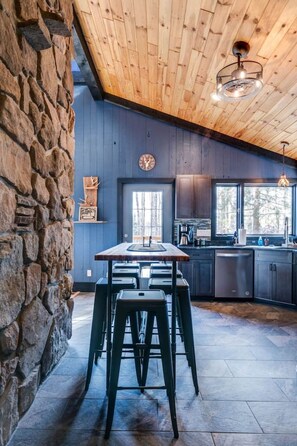 A modern kitchen with a stone wall, wooden ceiling, and a central island with stools. - Cozy Mountain Cabin, dark skies, and great views! (West Branch Township)