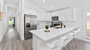 Una cocina moderna con armarios blancos, una isla central con taburetes y electrodomésticos como refrigerador y microondas.