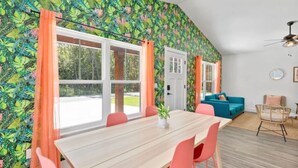 Un comedor con una mesa y sillas de madera, una planta en maceta y vistas al exterior a través de amplios ventanales con cortinas anaranjadas.