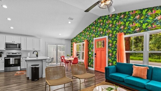 Una cocina moderna con armarios blancos y una isla central, un comedor con sillas rojas, y una sala de estar con un sofá color turquesa y una mesa de centro.