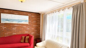 Ein Zimmer mit einer roten Couch, einem cremefarbenen Sessel, einer Ziegelwand und einem großen Fenster mit durchsichtigen Vorhängen.
