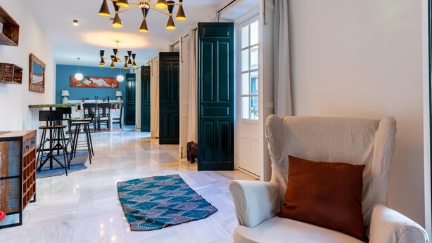 A spacious living area with a large armchair, a dining table with chairs, a kitchen in the background, and a patterned rug on the floor. - Holi-Rent Peñuelas (Sevilla)