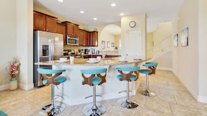 Una cocina moderna con una isla central, taburetes de bar, armarios de madera y electrodomésticos de acero inoxidable, y una escalera que sube a otro nivel.