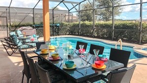Un área de piscina al aire libre con mesa de vidrio preparada para comer, rodeada de sillas y una piscina al fondo.