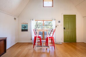 Una habitación con piso de madera, una puerta verde, una ventana con cortinas y una mesa con sillas rojas.