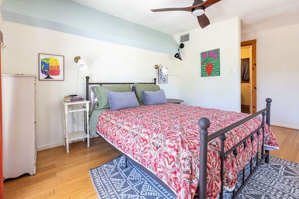 A bedroom with a bed, a bedside table, a lamp, a ceiling fan, and a door leading to another room.