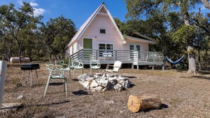 A pink cabin with a porch, surrounded by trees and outdoor furniture. - The Bohemian - A Hill Country Cottage (San Marcos)