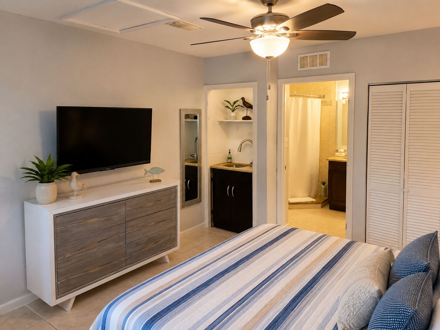 A bedroom with a bed, a TV, a ceiling fan, a dresser, a bathroom with a shower, and a closet.