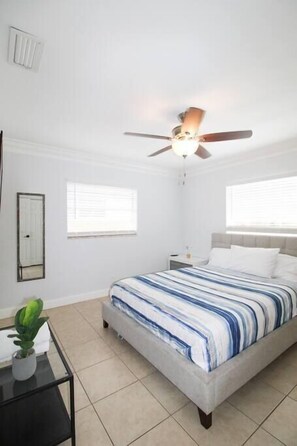 A bedroom with a bed, a ceiling fan, a nightstand, a potted plant, and a window.
