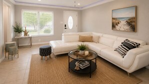Un salon avec un canapé d’angle blanc, une table basse en verre, un ventilateur de plafond et un tableau sur le thème de la plage.