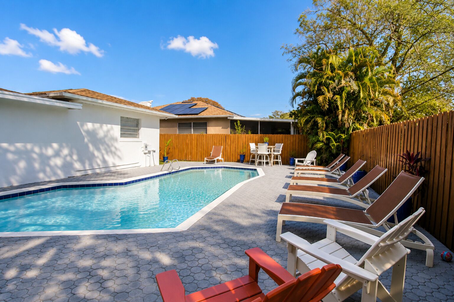 A swimming pool with lounge chairs and a table, surrounded by a wooden fence and greenery.