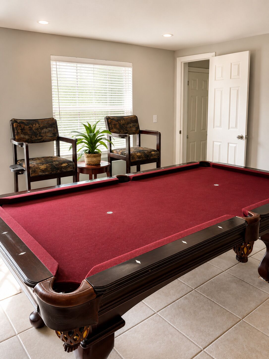 A pool table with a red felt surface, two wooden chairs with patterned cushions, a potted plant, and a white door in a room with beige walls and tiled flooring.