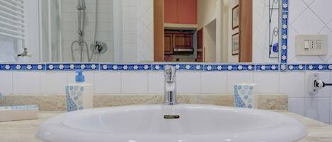 Une salle de bain avec un lavabo blanc, des carreaux bleus et blancs à motifs, une douche en verre et une vue sur une partie de la cuisine.