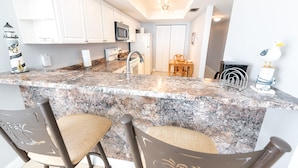 A kitchen with white cabinets, a granite countertop, a microwave, a refrigerator, and a dining area with a table and chairs. - Oceanfront 2BR Condo 6th Floor Corner Unit Indian Harbour Beach, 601 (Indian Harbour Beach)