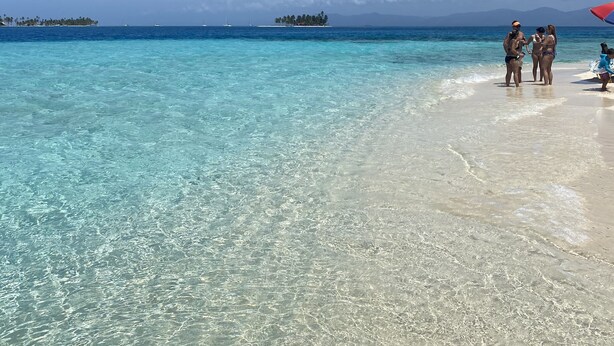 Una playa con aguas turquesas cristalinas, una orilla arenosa y islas a lo lejos.