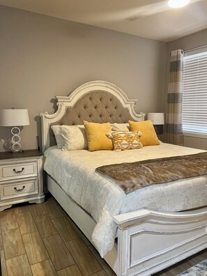 A bedroom with a tufted headboard, a large bed with patterned pillows, a nightstand with a lamp, and a window with blinds.