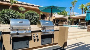 Un área de parrilla al aire libre con dos barbacoas, una piscina y un espacio de estar cubierto con sombrillas.