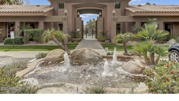 Un edificio con un arco central, un jardín ajardinado con fuente de agua y un cielo despejado.