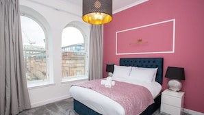 A hotel room with a large bed, a nightstand, a lamp, and a view of the city through large windows.