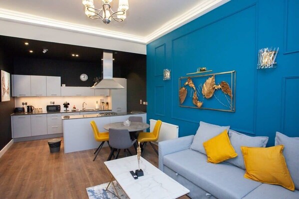 Una cocina moderna con armarios blancos, un comedor con una mesa redonda y sillas, y una sala de estar con sofá y cojines.