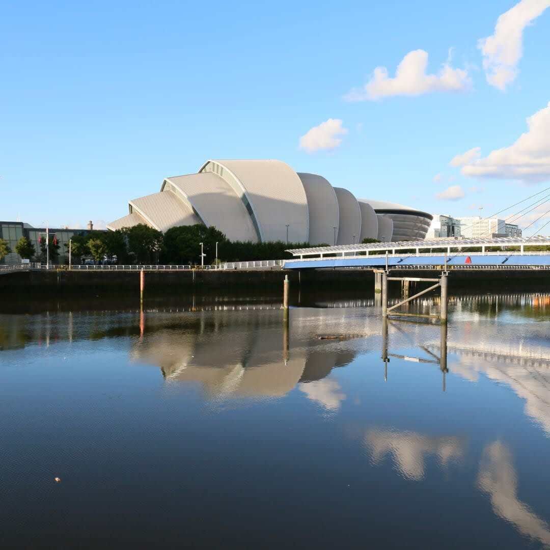 En moderne bygning med et unikt, buet design, en bro og et roligt vandspejl, der reflekterer bygningen.