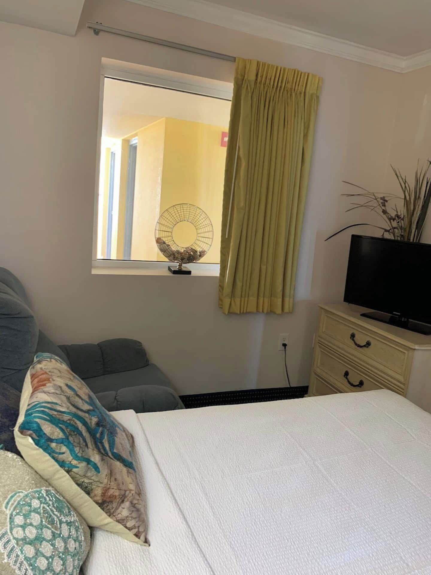 A bedroom with a bed, a dresser, a television, a window with curtains, and a fan on the windowsill.