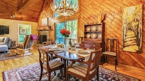 Ein rustikales Holzinterieur mit einem für vier Personen gedeckten Esstisch, umgeben von Holzstühlen. Eine Chandelüster hängt von der Decke, und im Hintergrund ist ein Kamin zu sehen.