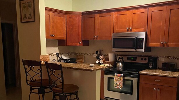 A compact kitchen with wooden cabinets, a microwave, an oven, and a coffee maker. There are two bar stools and a sofa in the living area. - Close to Beach and Fish Fry (Nassau)